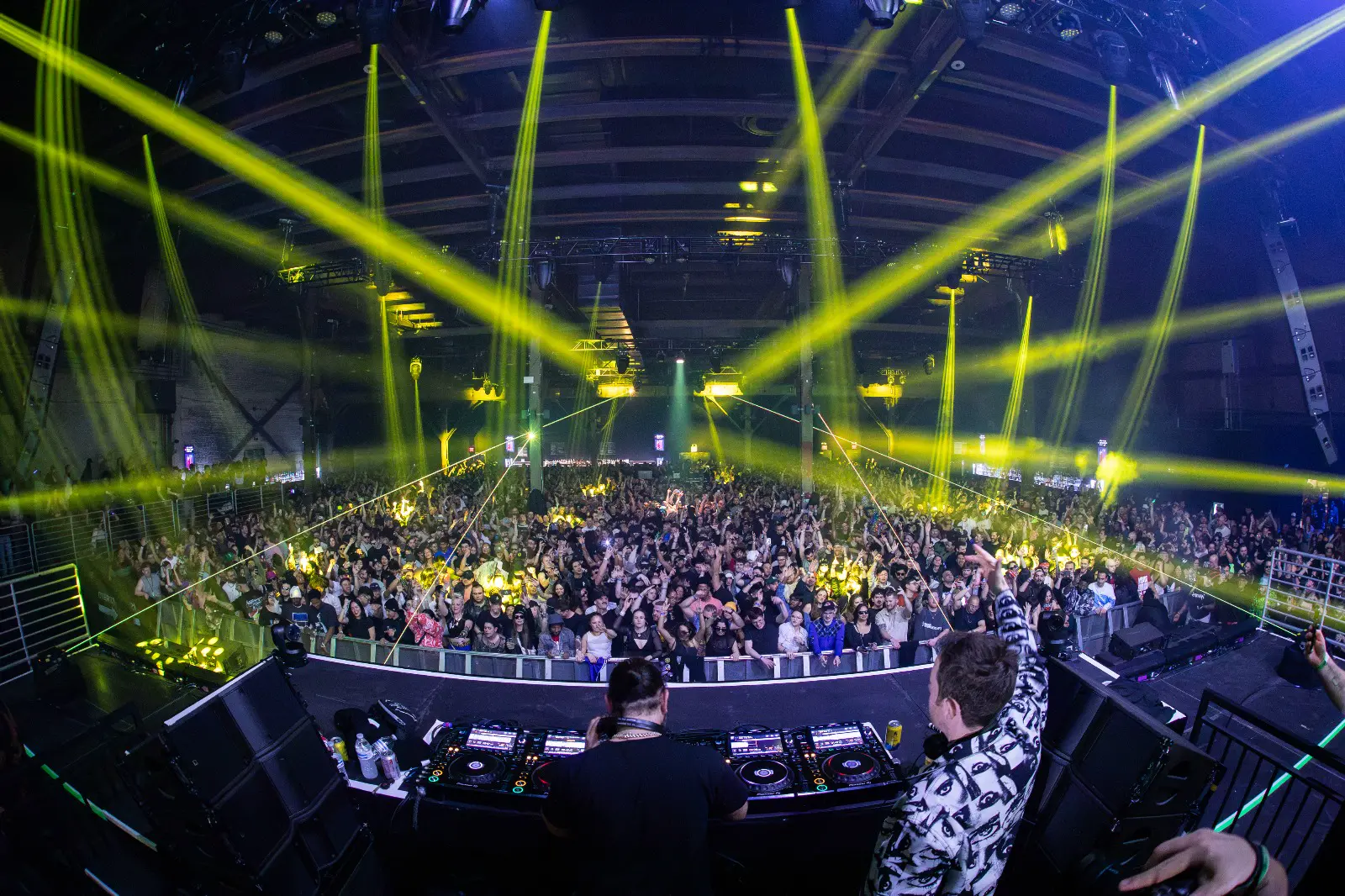 DJs on stage facing a packed room under yellow beam lights
